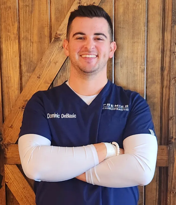 Dr. Dominic DeBlasie smiling in navy scrubs, arms crossed, in front of a barn wood wall