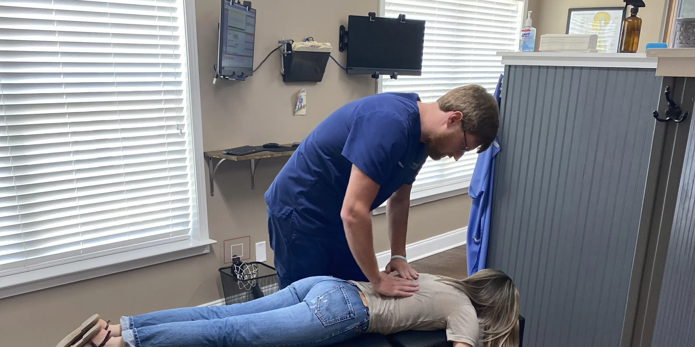 A doctor adjusting the lower back of a patient lying face down on an adjustment table, in a bright treatment room with window blinds
